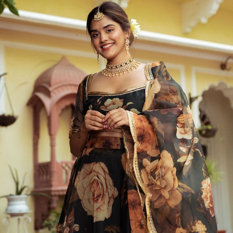 May include: A woman wearing a black and brown floral print lehenga with a gold trim. She is wearing a gold necklace and earrings. The background is a yellow wall.