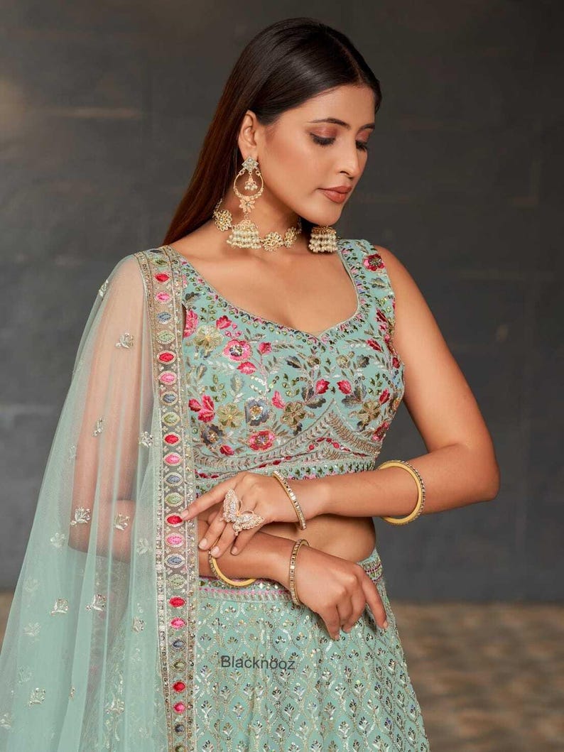 May include: A light turquoise embroidered formal dress with floral patterns and a matching sheer veil. The dress features gold bangles, a ring, and earrings. The text "Blacknboz" is visible on the dress.