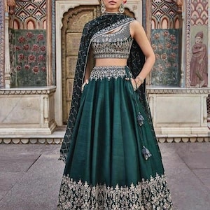Puede incluir: Un vestido de noche verde oscuro con intrincados bordados dorados. La parte superior sin mangas y la falda est&aacute;n adornadas con patrones detallados. Un chal a juego est&aacute; drapeado sobre los hombros. El vestido presenta un cuello alto y una falda fluida.