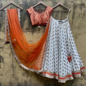May include: A white skirt with a gold pattern and orange trim. The skirt is paired with an orange and gold embroidered blouse.