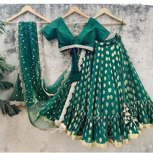 May include: A green and gold Indian Lehenga set, including a top, skirt, and scarf. The skirt and scarf have gold leaf patterns. The top has a V-neck and short sleeves. The set is displayed on a wooden hanger.