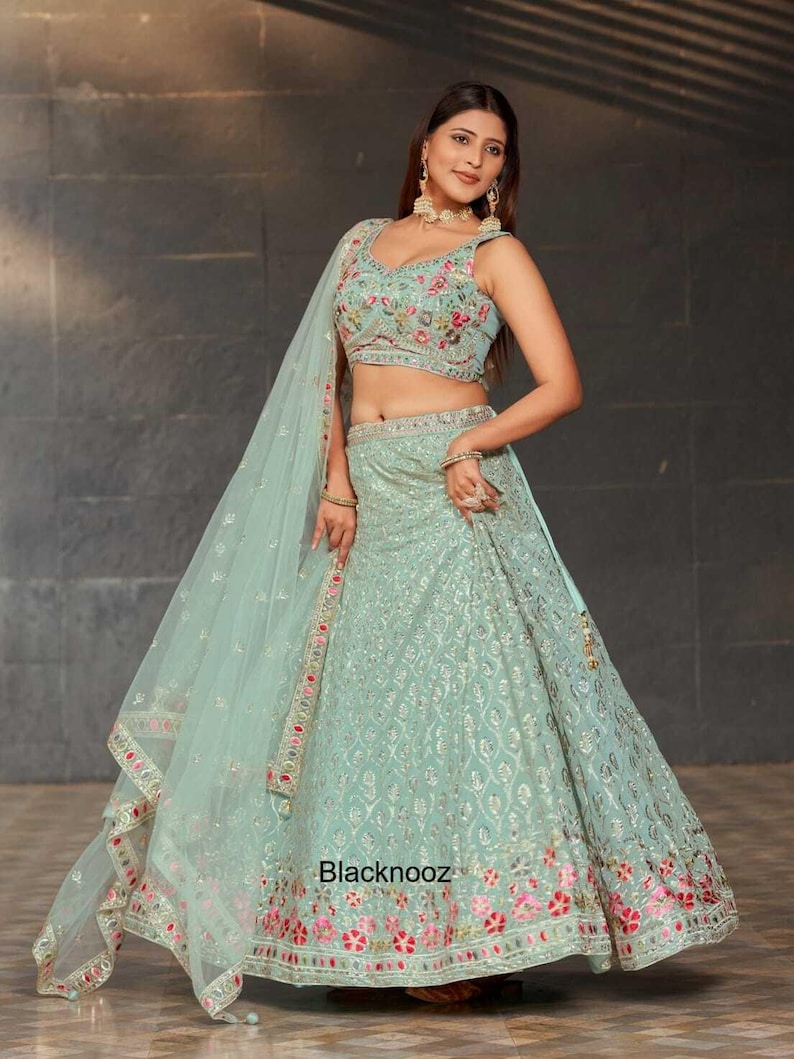 May include: A light green embroidered lehenga choli set. The sleeveless top features floral embroidery, and the skirt has intricate patterns. A matching sheer dupatta with a floral border completes the ensemble. The text "Blacknooz" is visible on the skirt.