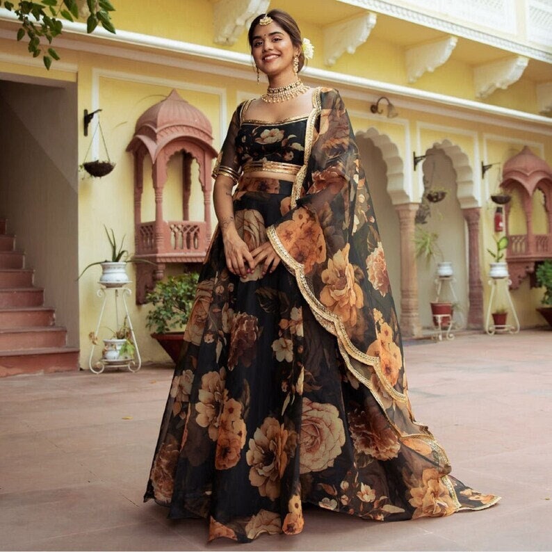 May include: A woman wearing a black and brown floral print lehenga with a gold embroidered border. She is wearing a matching black and brown floral print blouse with a gold embroidered border. She is also wearing a black sheer dupatta with gold embroidered trim.