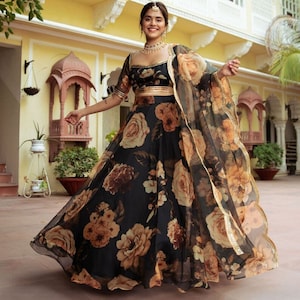 May include: A black and gold floral print lehenga with a sheer dupatta. The lehenga has a fitted bodice and a flowing skirt with a floral print. The dupatta is sheer and has a gold border.