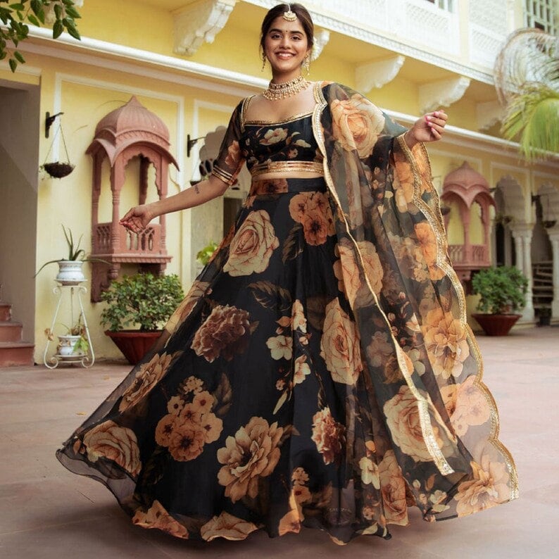 May include: A black and gold floral print lehenga with a sheer dupatta. The lehenga has a fitted bodice and a flowing skirt with a floral print. The dupatta is sheer and has a gold border.
