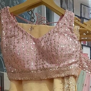 May include: A pink sleeveless blouse with a V-neckline and intricate gold embroidery. The blouse features a delicate floral pattern and a beaded trim.