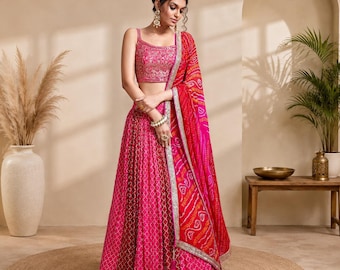 May include: A vibrant fuchsia lehenga set featuring a sleeveless blouse with intricate embroidery and a matching floor-length pleated skirt with a geometric pattern. A bright red dupatta with a traditional tie-dye print and silver border is draped.