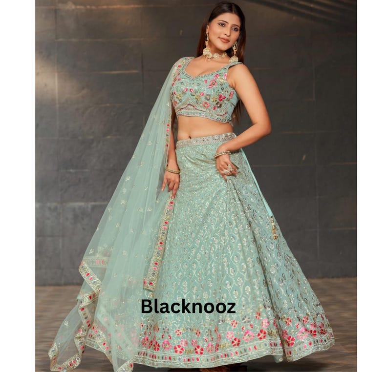 May include: A light green embroidered lehenga with a matching dupatta. The lehenga features floral embroidery in pink, red, and green. The top is sleeveless with a V-neckline and matching embroidery. The text "Blacknooz" is visible.