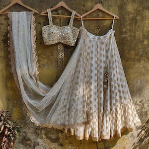 May include: A white and gold embroidered lehenga with a matching white and gold embroidered crop top. The lehenga has a white net dupatta with a gold border.