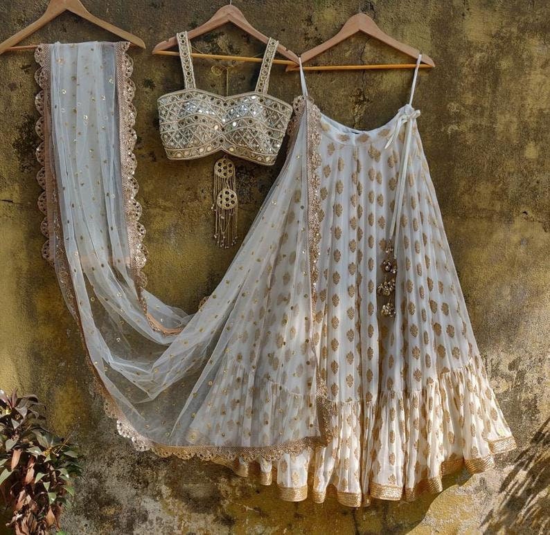 May include: A white and gold embroidered lehenga with a matching white and gold embroidered crop top. The lehenga has a white net dupatta with a gold border.