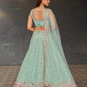 May include: A light green lehenga with intricate embroidery and a matching top. The outfit features a sheer dupatta and is adorned with pink and silver accents. The text "Blacknooz" is visible at the bottom.