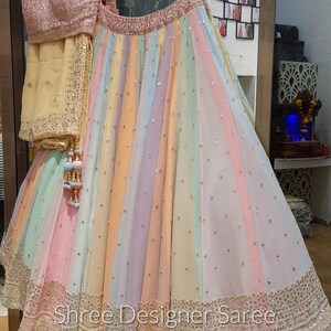 May include: A rainbow-coloured skirt with a gold embroidered border and a matching pink blouse. The skirt is made of a sheer fabric and has a flowing silhouette. The text "Shree Designer Saree" is visible at the bottom of the skirt.