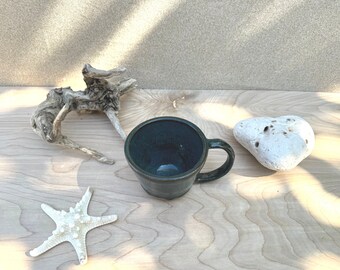 Pode incluir: Uma caneca de cerâmica verde escura com alça, uma estrela do mar branca, um pedaço de madeira flutuante e uma pedra branca com buracos em uma superfície de madeira clara.