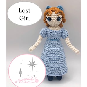 May include: Crocheted doll with brown hair, blue eyes, and a light blue dress. The doll is standing on a white background with a star design and the text "Lost Girl".