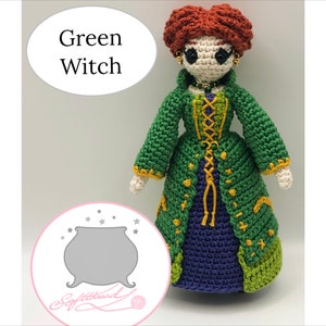 Pode incluir: Boneca de bruxa verde de crochê com um vestido roxo, detalhes amarelos e um caldeirão no fundo. A boneca tem cabelo ruivo e olhos castanhos.