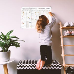 May include: A large white wall calendar featuring a grid layout with the German text Rein ins Abenteuer at the top. The calendar includes various small colored icons and markers distributed across the monthly columns for tracking activities.
