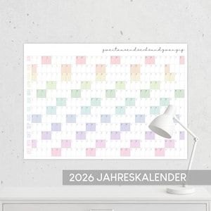 May include: A large 2026 yearly calendar with pastel-colored blocks for each month. The calendar is displayed on a white wall above a white desk. The text "2026 JAHRESKALENDER" is printed at the bottom of the calendar.