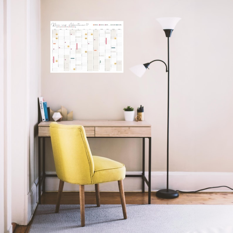 May include: A wall-mounted annual calendar featuring the text "Rein ins Abenteuer" at the top, displayed above a wooden desk with a yellow upholstered chair and a tall black floor lamp. The calendar grid includes columns for months and days.