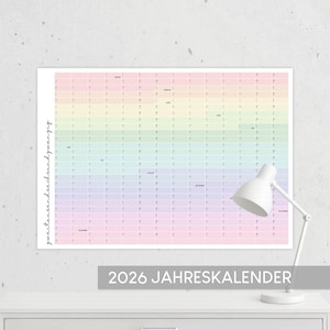 May include: A large, rectangular 2026 Jahreskalender calendar with a pastel rainbow gradient background. The calendar is displayed on a white surface, with a white desk lamp to the right. The text "2026 JAHRESKALENDER" is printed in a gray rectangle at the bottom.
