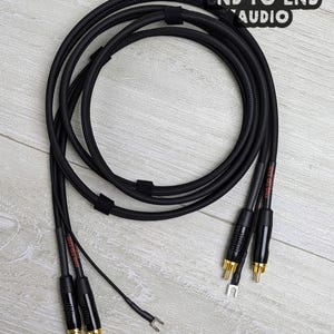 May include: A black braided audio cable coiled in a circle, featuring gold-plated RCA connectors and spade lugs at the ends. The cable includes black heat-shrink tubing with the text END-TO-END AUDIO printed in red on the connector housings.