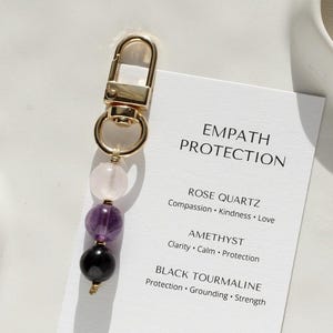 May include: A gold-tone keychain with a lobster clasp, strung with rose quartz, amethyst, and black tourmaline beads. A white card displays "EMPATH PROTECTION" and stone properties. The website address is also visible.