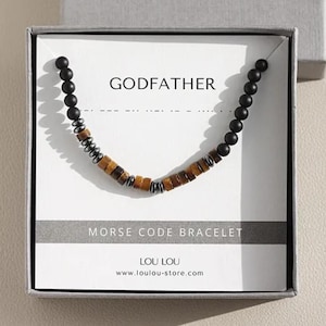 Op de afbeelding: Een kralenarmband met het woord "GODFATHER" in morsecode, gepresenteerd in een grijze geschenkverpakking. De armband heeft zwarte, bruine en zilveren kralen. De doos is voorzien van de merknaam "LOU LOU".
