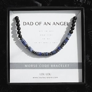 May include: A gift box containing a Morse code bracelet. The bracelet features blue and black beads. The card inside the box reads "DAD OF AN ANGEL" and "MORSE CODE BRACELET". The box is filled with black shredded paper.