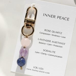 May include: A gold-coloured keychain featuring three gemstone beads: rose quartz, lavender amethyst, and sodalite. A white card with the text "INNER PEACE" and gemstone properties is attached. The website "loulou-store.com" is also visible.