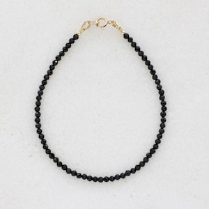 Könnte beinhalten: Zartes Armband mit schwarzen Perlen und einem goldfarbenen Verschluss. Das Armband besteht aus kleinen, runden, schwarzen Perlen. Der Verschluss ist ein einfacher Haken- und Ösenverschluss. Das Armband liegt auf einer weißen Oberfläche.
