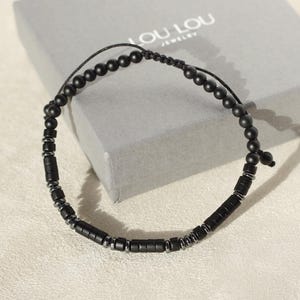 May include: Black beaded bracelet featuring a combination of round and rectangular beads. The bracelet is displayed on a light surface, with a grey box in the background that reads "LOU LOU JEWELRY". The bracelet is adjustable.