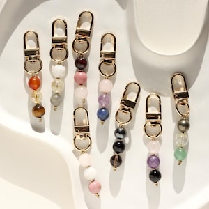 Könnte beinhalten: Goldfarbene Schlüsselanhänger mit Strängen aus Edelsteinperlen in verschiedenen Farben. Die Perlen sind in Orange, Gelb, Weiß, Rosa, Lila, Blau, Schwarz und Grün gehalten. Die Schlüsselanhänger sind auf einer weißen, geschwungenen Oberfläche angeordnet.