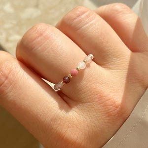 Può includere: Un delicato anello di perline con una fila di piccole pietre preziose colorate. L'anello presenta una combinazione di perline rosa, oro e rosso scuro, creando un accessorio delicato ed elegante. Un gioiello semplice ma raffinato.