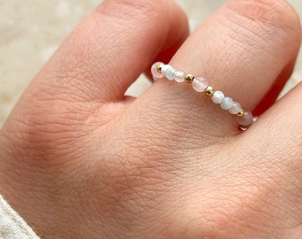 May include: A beaded ring featuring small, round, pale blue and pink beads interspersed with gold-colored beads. The ring is worn on a finger, showcasing the delicate design and color palette. The ring is a piece of jewelry.
