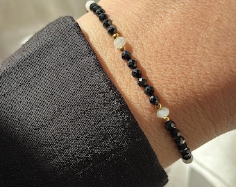 May include: A delicate beaded bracelet featuring small, faceted black beads arranged in a continuous line, interspersed with two translucent, irregular white stones and tiny gold-toned metal spacers, worn on a wrist against a black sleeve.