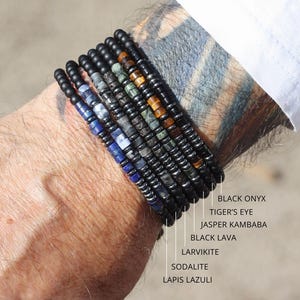 May include: A selection of gemstone bracelets, including Black Onyx, Tiger's Eye, and Lapis Lazuli, are displayed on a wrist. The bracelets feature black beads and various colored gemstones. The text "CHOOSE YOUR GEMSTONE" is visible.
