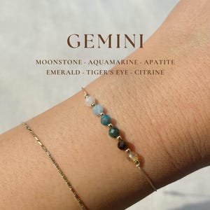 May include: A gold bracelet with a variety of gemstones, including moonstone, aquamarine, apatite, emerald, tiger's eye, and citrine. The bracelet is on a person's wrist. GEMINI is written in large letters at the top of the image.  The text below GEMINI reads: MOONSTONE. AQUAMARINE. APATITE. EMERALD. TIGER'S EYE. CITRINE.