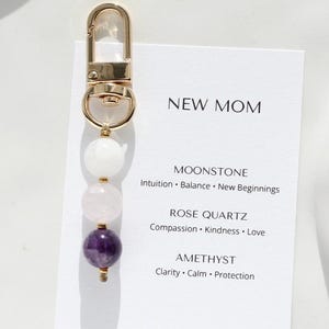 Könnte beinhalten: Ein goldfarbener Schlüsselanhänger mit einem Anhänger aus weißen, rosa und lila Edelsteinen. Der Schlüsselanhänger wird mit einer weißen Karte mit dem Text "NEW MOM" und Edelsteinbeschreibungen präsentiert.