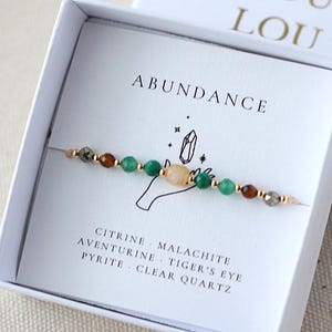 May include: A beaded bracelet featuring citrine, malachite, aventurine, tiger's eye, pyrite, and clear quartz beads on a string. The bracelet is presented in a white box labelled ABUNDANCE with the brand name LOU LOU printed on the lid.