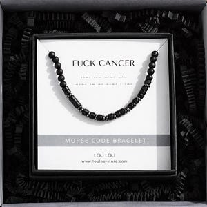 May include: A black beaded Morse code bracelet displayed on a white card inside a black gift box. The card features the text FUCK CANCER, MORSE CODE BRACELET, and LOU LOU www.loulou-store.com. The bracelet consists of matte black round beads and small metallic spacers.