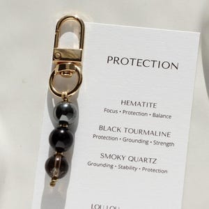 Può includere: Un portachiavi color oro con tre perline di pietre preziose: ematite, tormalina nera e quarzo fumé. È attaccato un biglietto bianco con la parola "PROTECTION" e le proprietà delle pietre preziose. È visibile anche il sito web www.loulou-store.com.