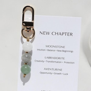 May include: A gold-toned metal keychain featuring three spherical beads in white, grey, and green, attached to a display card labeled NEW CHAPTER with text detailing Moonstone, Labradorite, and Aventurine properties and the website loulou-store.com.