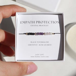 Könnte beinhalten: Ein schwarzes Armband mit schwarzen Turmalin-, Amethyst- und Rosenquarzperlen. Das Armband wird in einer weißen Schachtel mit dem Text "EMPATH PROTECTION CRYSTAL BRACELET" präsentiert. Die Schachtel listet auch die Kristallarten und den Markennamen auf.