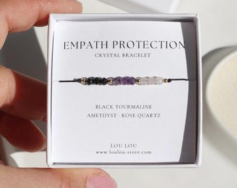 Puede incluir: Una pulsera de cordón negro con cuentas de turmalina negra, amatista y cuarzo rosa. La pulsera se muestra en una caja blanca con el texto "EMPATH PROTECTION CRYSTAL BRACELET". La caja también enumera los tipos de cristales y el nombre de la marca.