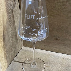 Könnte beinhalten: Ein klares Weinglas aus Glas mit den Worten "FLUT" und "EBBE" auf der Seite graviert. Das Glas hat einen langen Stiel und einen breiten Boden.