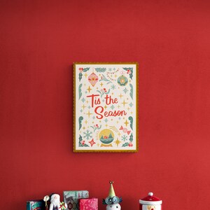 May include: A framed print with a gold border and a white background. The print features a festive design with ornaments, snowflakes, and the text "Tis the Season". The print is hanging on a red wall above a shelf with various Christmas decorations.
