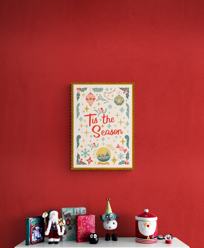 May include: A framed print with a gold border and a white background. The print features a festive design with ornaments, snowflakes, and the text "Tis the Season". The print is hanging on a red wall above a shelf with various Christmas decorations.