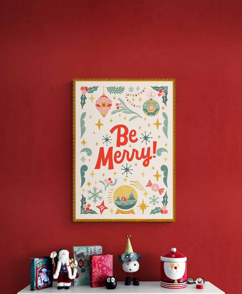 Pu&ograve; includere: Una stampa incorniciata con sfondo bianco e cornice dorata. La stampa presenta un design festivo con un globo di neve, stelle e il testo "Be Merry!" in lettere rosse e bianche.