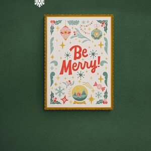 Pu&ograve; includere: Una stampa incorniciata con sfondo bianco e cornice dorata. La stampa presenta un design festivo con una sfera di neve, ornamenti, stelle e il testo "Be Merry!" in rosso.