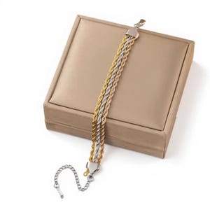 Könnte beinhalten: Ein dreifarbiges Seilarmband mit Gold-, Silber- und Roségoldtönen. Das Armband wird auf einer beigen Schmuckschatulle präsentiert. Es hat eine silberne Kettenverlängerung und einen Verschluss.