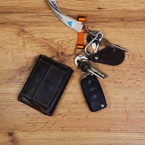 May include: A black leather wallet, keys, and a car key fob rest on a wooden surface. A lanyard with a white strap and blue heart design is attached to a metal clip holding the keys. The wallet is dark brown with visible stitching.